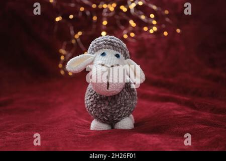 Niedlicher Plüsch-Widder zur Gestaltung der Weihnachtskarte. Grau und weiß handgemachte Amigurumi Schafe gehäkelt mit blauen Augen steht auf rot weiche Decke gegen Rücken Stockfoto