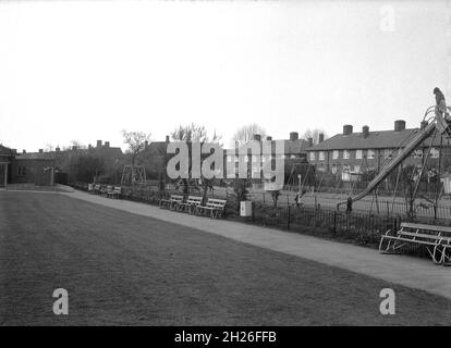 Historischer Blick auf einen öffentlichen Vorstadtpark mit Sportplatz in den 1950er-Jahren, der sich an einer Reihe von lokalen rathäusern befindet, England, Großbritannien. Kinder können neben dem Park auf einem Spielplatz mit traditioneller Ausrüstung wie einer Metallrutsche, einer Hexenhütte und einem Kreisverkehr, Maybe Abbey Wood oder Eltham in South London, spielen sehen. Stockfoto