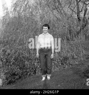 Die 60er Jahre, historisch, eine junge Frau, die auf einem Feld steht und ein kurzärmeliges Oberteil und eine Demin-Jeans mit Umschlagboden trägt. Diese wurden in den späten 1950er und frühen 60er Jahren modisch und der coole Look, als Rockabilly populär wurde. Früher waren Kleider und Röcke die Norm für Frauen jeden Alters und nicht Hosen oder Hosen und Demin waren wirklich nur für die Handarbeit getragen worden. Stockfoto