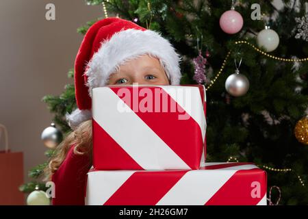 Ein kleines Mädchen in einem Weihnachtsmannhut versteckt sich hinter einem Stapel von Neujahrsschachteln mit Geschenken. Nahaufnahme eines lustigen Kindes auf dem Hintergrund eines Weihnachtsfestes Stockfoto