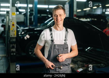 Mittellange Aufnahme eines lächelnden, hübschen jungen Mechanikers mit Uniform und speziellem Schlüsselschlüssel Stockfoto