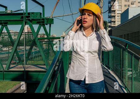 Schöne junge Ingenieur oder Supervisor, trägt Schutzhelm, Gespräch am Telefon mit ihrem Vorgesetzten während der Inspektion einer Eisenbahn Fußgängerbrücke. c Stockfoto