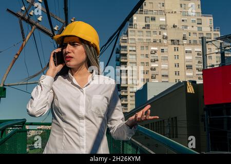 Schöne junge Ingenieur oder Supervisor, trägt Schutzhelm, Gespräch am Telefon mit ihrem Vorgesetzten während der Inspektion einer Eisenbahn Fußgängerbrücke. c Stockfoto