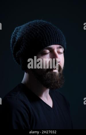 Authentisches dramatisches Porträt eines Millennials mit einem Bart in einem schwarzen Strickmütze auf dunklem Hintergrund Stockfoto
