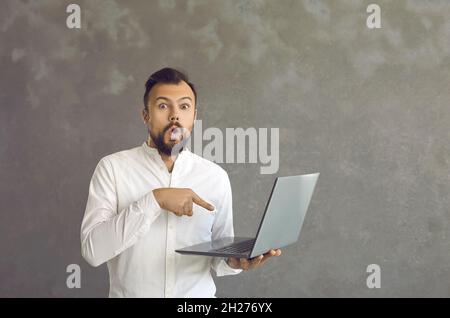 Schockierter Mann zeigt mit einem Finger auf seinen Laptop, den er auf grauem Hintergrund in der Hand hält. Stockfoto