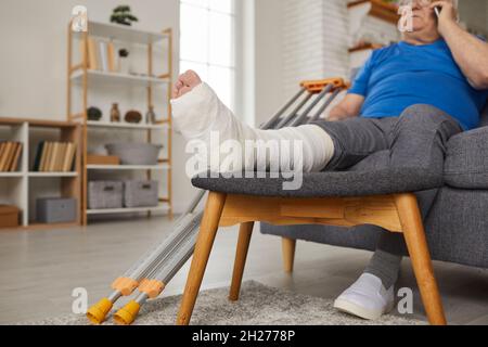 Älterer Mann mit gebrochenem Bein in Gipsabguss sitzt auf der Couch und spricht am Telefon Stockfoto