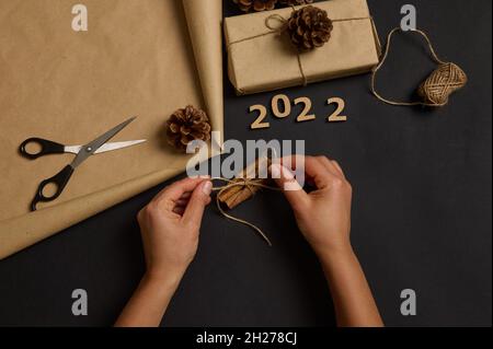 Vorbereitung auf Weihnachten. Frauenhände binden Zimt mit einem Seil. Einfaches, minimalistisches Geschenk, eingewickelt in braunem Bastelpapier, verziert mit Kieferkegel, woo Stockfoto