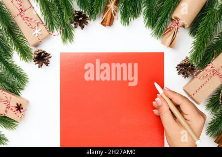 Konzept des Schreibens an den Weihnachtsmann und der Wünsche. Frau Hand auf rotem Papier und Hintergrund von verzierten Tannenzweigen Stockfoto
