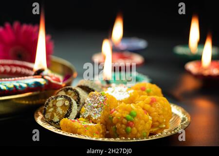 Köstliche Mithai Laddu, Meetha Laddoo Burfi, Anjeer Roll In Brass Thali Und Diya Deep Oder Dia Beleuchtet Auf Schwarzem Hintergrund. Thema Für Diwali Pooja, Navratri Stockfoto