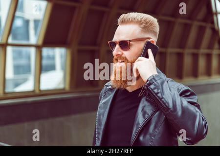 Der bärtige rothaarige Mann mit Sonnenbrille und Lederjacke führt Telefongespräche. Stockfoto