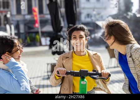 Fröhliche junge Gesellschaft von lächelnden multirassischen Freunden in der Stadt zu sprechen, Freundinnen Spaß haben Zeit zusammen, bunte Sommer Hipster Mode-Stil, Reiten auf Elektroroller, moderne Jugend. Hochwertige Fotos Stockfoto