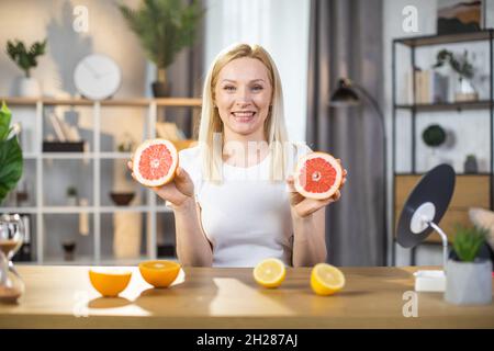 Porträt einer angenehmen Frau, die Grapefruitscheiben hält, während sie mit aufrichtigem Lächeln auf dem Gesicht am Tisch sitzt. Kaukasische Blondine in weißem T-Shirt, die zu Hause Schönheitsrituale genießt. Stockfoto