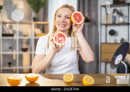 Porträt einer angenehmen Frau im Alter von 40 Jahren, die Grapefruitscheiben hält, während sie mit einem aufrichtigen Lächeln auf dem Gesicht am Tisch sitzt. Kaukasische Blondine in weißem T-Shirt, die zu Hause Schönheitsrituale genießt. Stockfoto