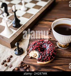 Schachfiguren auf dem Schachbrett während der Mittagspause. Entspannende Logikspiele am Morgen mit einer Tasse Kaffee und süßem Donut. Quadratisch abgeschnitten Stockfoto