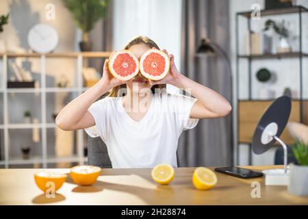 Portrait einer hübschen Teenagerin, die Grapefruitscheiben über die Augen hält, während sie am Tisch sitzt. Kaukasische junge Frau mit perfekter frischer Haut posiert drinnen. Stockfoto