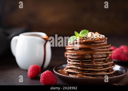 Stapel von Schokoladenpfannkuchen mit Sauce und gehackten Nüssen, serviert mit frischen Himbeeren und Minzblättern. Süße Pfannkuchen zum Frühstück Stockfoto