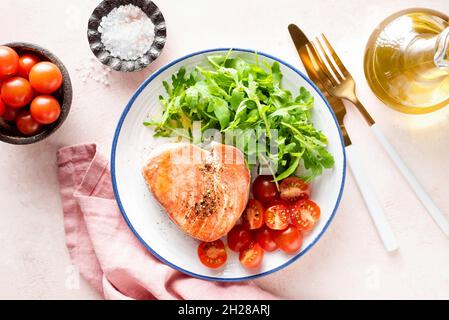 Gebratenes rosafarbenes Thunfischsteak mit Rucola und Tomaten auf einem Teller, rosa Hintergrund, Draufsicht. Gesunde Mahlzeit reich an Omega 3 Stockfoto