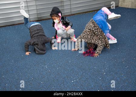 Bildung Vorschule 4-5 Yar alt zwei Mädchen und ein Junge versuchen, Handstände oder Kopf steht draußen auf dem Spielplatz zu tun, tragen Gesichtsmasken Covid-19 Stockfoto