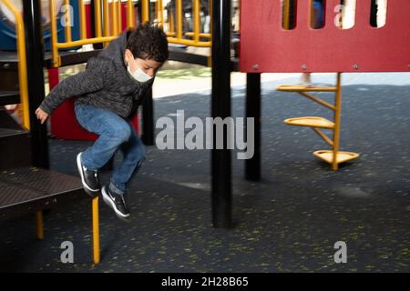 Bildung Vorschule 4-5-Jährige im Freien auf dem Spielplatz Junge springt von Spielgeräten, trägt Gesichtsmaske, um vor Covid-19-Infektion zu schützen Stockfoto