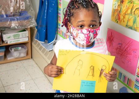 Bildung Preschool Mädchen im Alter von 4-5 Jahren, das die Zeichnung ihrer Familie hochhält und eine Gesichtsmaske trägt, um sich vor einer Covid-19-Infektion zu schützen Stockfoto