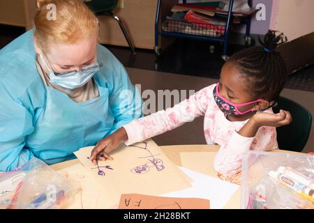 Bildung Preschool Mädchen im Alter von 4-5 Jahren, das der Lehrerin über ihre Zeichnung erzählt, die beschreibt, was auf dem Bild zu sehen ist, beide tragen Gesichtsmasken Covid-19 Stockfoto