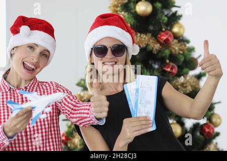 Zwei lächelnde Frauen mit weihnachtsmannmützen halten Tickets und Daumen hoch aus der Nähe Stockfoto