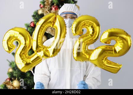 Arzt in Schutzanzug, Maske und Brille hält die Zahlen 2022 vor dem Hintergrund des Neujahrsbaums Stockfoto