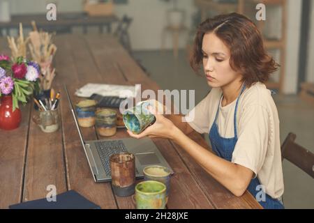 Seriöse junge kaukasische Frau sitzt am Holztisch mit Laptop und untersucht Keramik-Becher in Töpferwerkstatt Stockfoto