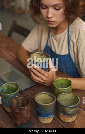 Konzentrierte junge Frau, die mit einem Laptop am Holztisch sitzt und handgefertigte Becher untersucht, bevor sie Käufer schickt Stockfoto