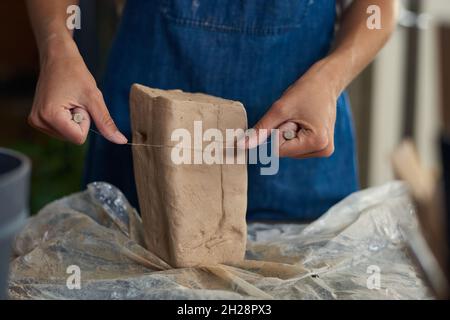 Nahaufnahme eines nicht erkennbaren Töpfers in einer Jeanschürze, die Ton mit abgeschnittenem Draht in der Werkstatt schneidet Stockfoto