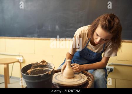 Konzentrierte junge kaukasische Frau in Denim-Schürze, die am Töpferrad sitzt und in der Werkstatt eine Tonvase modelliert Stockfoto