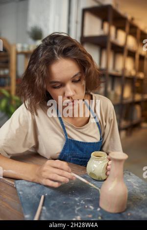 Konzentrierter junger kaukasischer Töpfer, der am Tisch sitzt und Vase malt, während er in der Werkstatt an seinem Design arbeitet Stockfoto