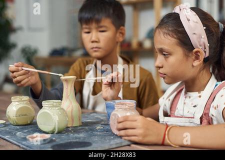 Konzentrierte, multiethnische Schulkinder, die am Tisch sitzen und mit Pinsel arbeiten, während sie im Unterricht Vasengestaltung entwerfen Stockfoto
