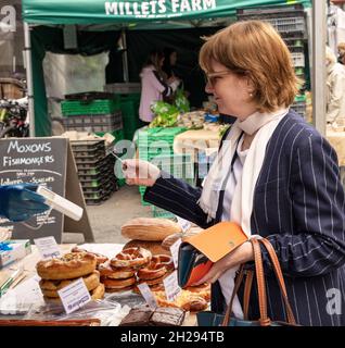 Elegant gekleidete Frau in einer Jacke mit Kreidestreifen, die Lebensmittel auf einem Markt im Freien in Bute St, South Kensington, London, kauft Stockfoto