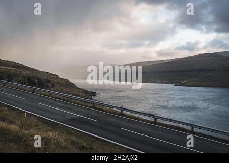 Leere Straße umgeben von skandinavischer Natur, braunen Feldern und Bergen, launisch dramatischer Sonnenuntergang Himmel Stockfoto