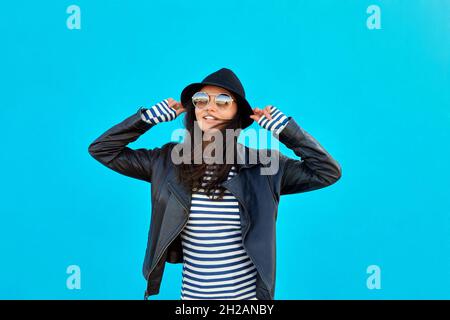 Entzückte trendige Frau in schwarzer Lederjacke und Sonnenbrille berühren Hut blauen Hintergrund Stockfoto