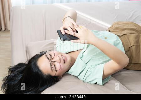 Schöne Brille Asiatische Frau liegt auf dem Rücken, plaudert, tippt und schreibt mobil in ihrem Schlafzimmer mit einem pastellgrün-braunen Farbthema. Stockfoto