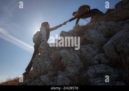 Hilfe- und Hilfekonzept. Silhouetten von zwei Menschen, die Berge besteigen und gegen den blauen Himmel helfen. Gehen Sie in den Wolken. Discovery Travel Direction Konzept Stockfoto