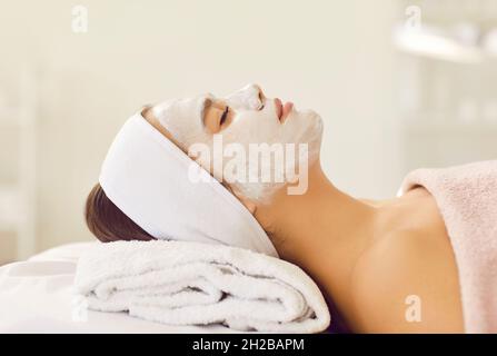 Schöne Frau mit Stirnband und reinigende weiße Maske auf ihrem Gesicht entspannt im Spa oder Schönheitssalon. Stockfoto