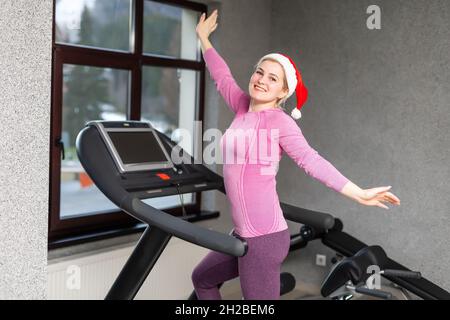 Ein Mädchen in einem Weihnachtsmannhut macht Fitnessübungen im Fitnessstudio, trägt Sportuniform, Weihnachten Fitness-Aktivität. Neujahr. Weihnachten, Urlaub, Fitness Stockfoto