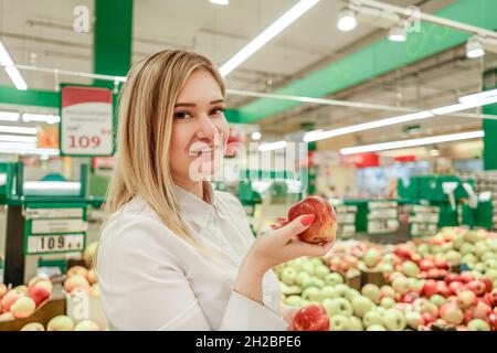 Schöne junge lächelnde Geschäftsfrau nimmt Äpfel, während sie in einem Lebensmittelgeschäft einkauft. Nahaufnahme Porträt mit Apfel in der Hand. Stockfoto
