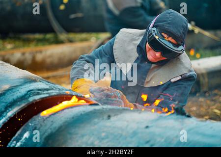 Der Schweißer schneidet große Metallrohre mit Oketylenschweißen. Ein Arbeiter auf der Straße schneidet tagsüber Rohre mit großem Durchmesser und Funken und Feuer fliegen. Stockfoto