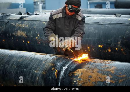 Der Schweißer schneidet große Metallrohre mit Oketylenschweißen. Ein Arbeiter auf der Straße schneidet tagsüber Rohre mit großem Durchmesser und Funken und Feuer fliegen. Stockfoto