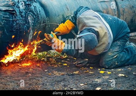 Der Schweißer arbeitet tagsüber mit Acetylenschweißen im Freien. Ein Arbeiter in Schutzkleidung schneidet ein Metallrohr zur Vergasung. Reparatur und Wartung der Gaspipeline. Stockfoto