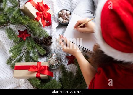 Draufsicht Frau im weihnachtsmütze schreibt Weihnachtswünsche in Notizbuch um Tannenzweige und Geschenke. Stockfoto