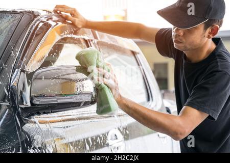 Autowascharbeiter mit T-Shirt und schwarzer Kappe reinigen das Auto in der Autowaschanlage mit einem Schwamm, Konzept für die Autopflege. Stockfoto