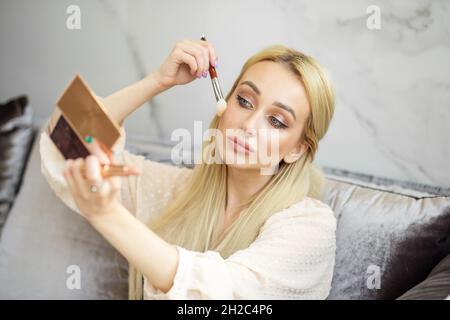 Junge blonde Frau trägt Kosmetikpulver auf ihr Gesicht. Kleiner Spiegel in der Hand. Das Konzept von Pflege und Make-up, Lifestyle und Schönheit. Stockfoto