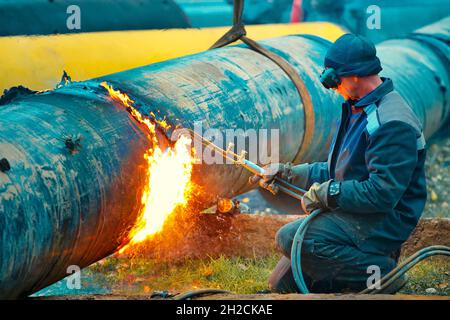 Der Schweißer schneidet große Metallrohre mit Oketylenschweißen. Ein Arbeiter auf der Straße schneidet tagsüber Rohre mit großem Durchmesser und Funken und Feuer fliegen. Stockfoto