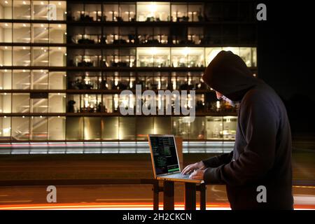 Symbolisches Bild Computerhacker, Computerkriminalität, Cyberkriminalität, Cyberangriff, Hacker mit Laptop vor einem Bürogebäude, hackt in Computersystem Stockfoto
