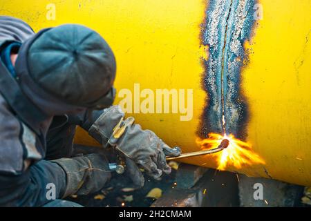 Der Schweißer schneidet ein großes gelbes Rohr mit Acetylenschweißen zur Vergasung. Entsorgung von alten, gebrauchten Metallrohren. Authentische Workflow-Szene. Industrieller Hintergrund. Stockfoto